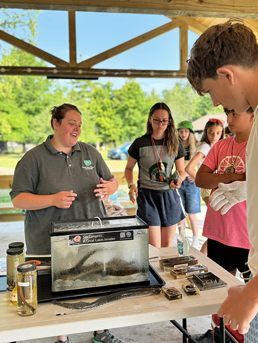 A woman in a Great Lakes Fishery Commission shirt stands behind a tank of live sea lamprey while a group of youth gather around.